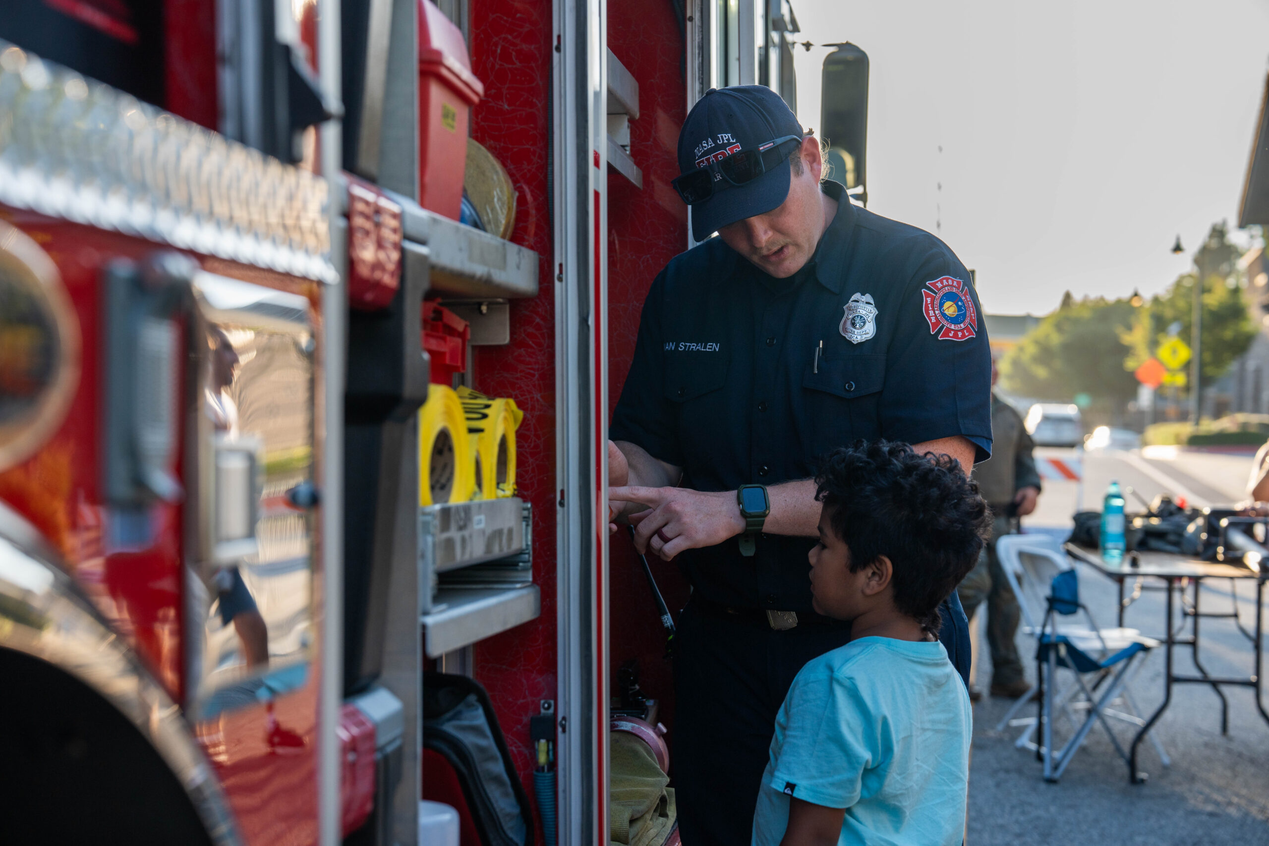 A firefighter in uniform explaining tools stored inside an open fire engine compartment to a young child.